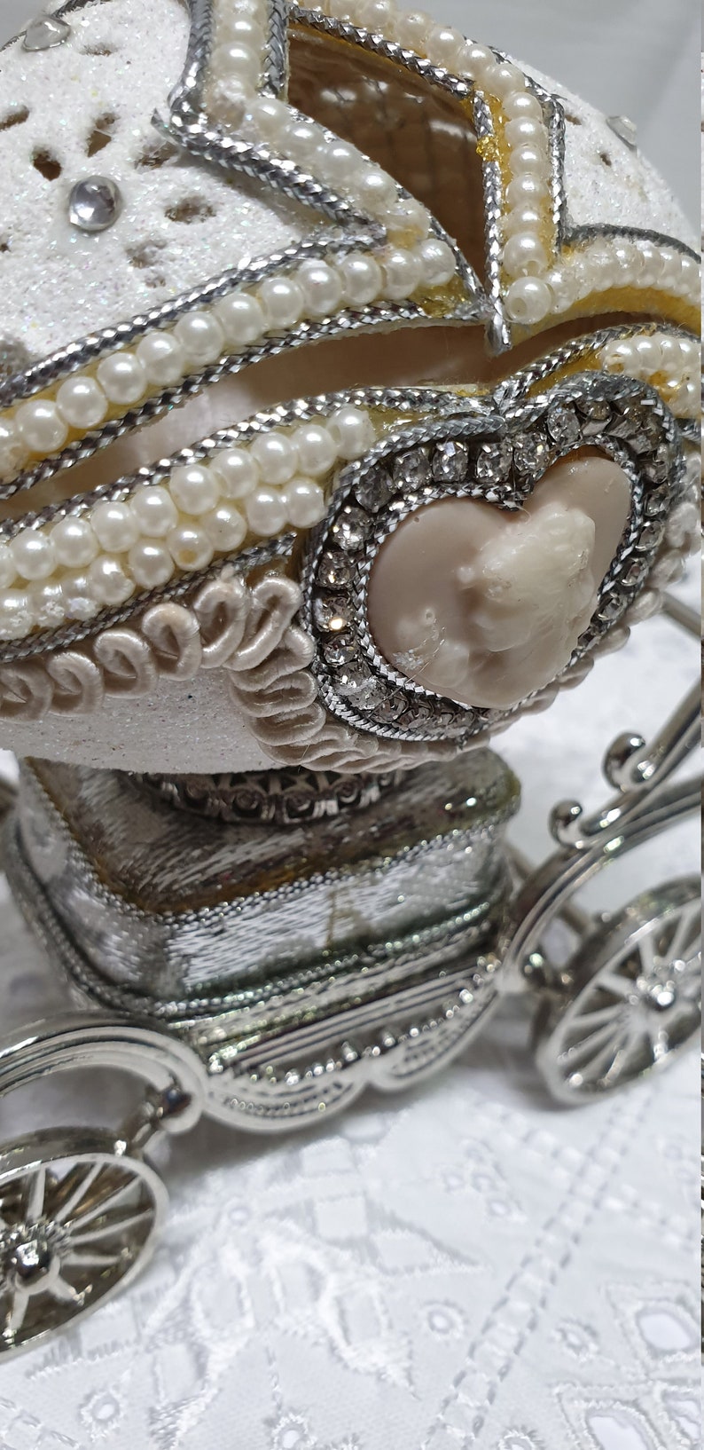 May include: A white porcelain egg decorated with pearls and rhinestones, set in a silver carriage with wheels. The egg has a heart-shaped opening with a cameo inside.