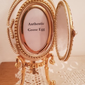May include: A white goose egg decorated with gold trim and pearls, with a hinged lid that opens to reveal a small interior space. The egg is sitting on a gold stand with three legs. The egg has a small picture frame with the text "Authentic Goose Egg" on it.