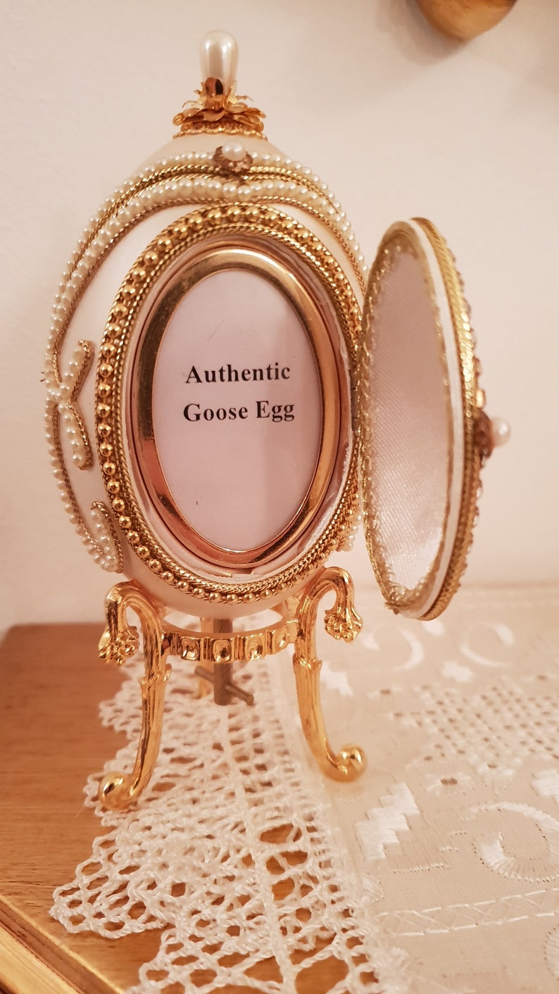 May include: A white goose egg decorated with gold trim and pearls, with a hinged lid that opens to reveal a small interior space. The egg is sitting on a gold stand with three legs. The egg has a small picture frame with the text "Authentic Goose Egg" on it.