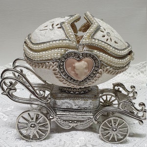 May include: A white porcelain egg with pearl accents and a silver carriage base. The egg is decorated with a pink cameo of a couple inside a heart.