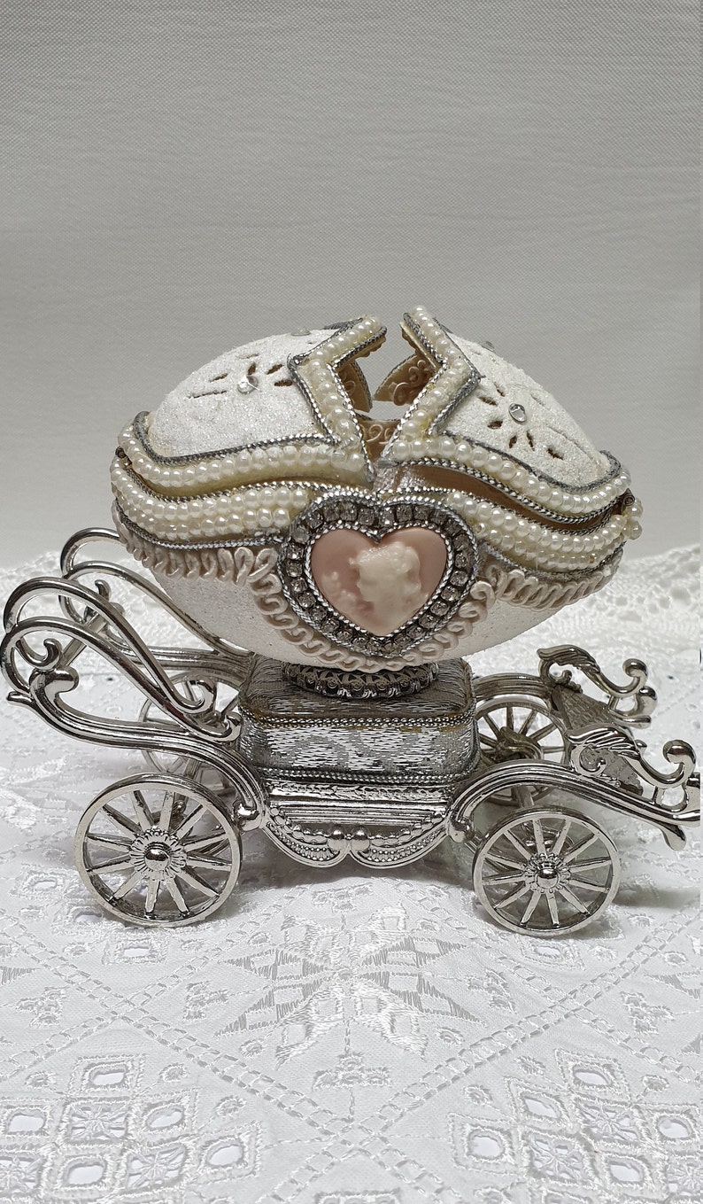 May include: A white porcelain egg with pearl accents and a silver carriage base. The egg is decorated with a pink cameo of a couple inside a heart.