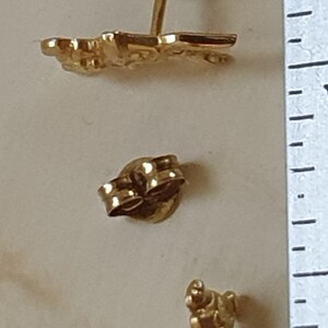 May include: A pair of gold-tone earrings featuring a stylized figure design. The earrings are made of metal and have a post back closure.