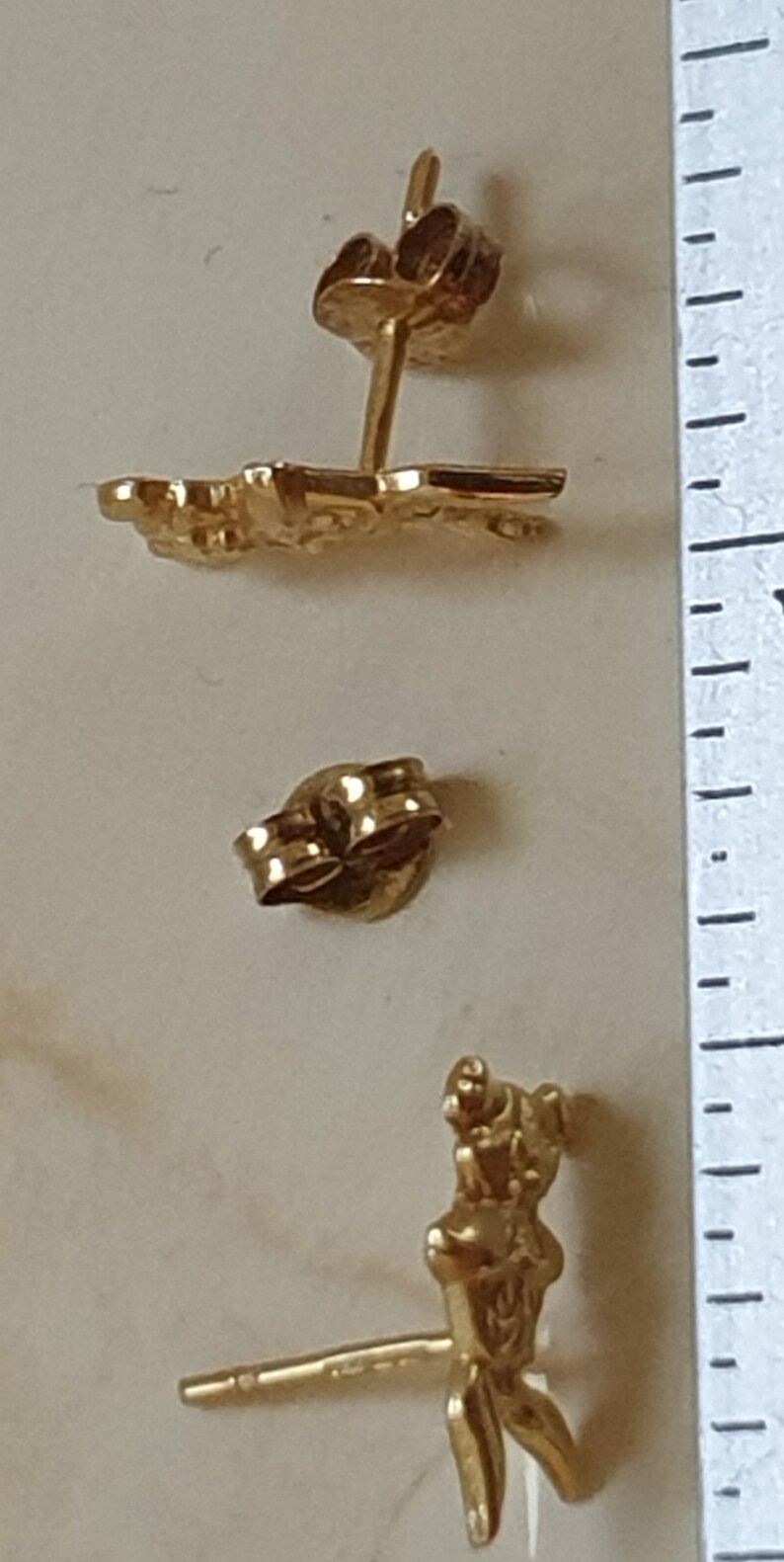 May include: A pair of gold-tone earrings featuring a stylized figure design. The earrings are made of metal and have a post back closure.