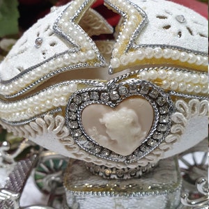May include: A white porcelain egg decorated with pearls and rhinestones, shaped like a carriage with a heart-shaped cameo on the front. The carriage has silver wheels and a silver base.