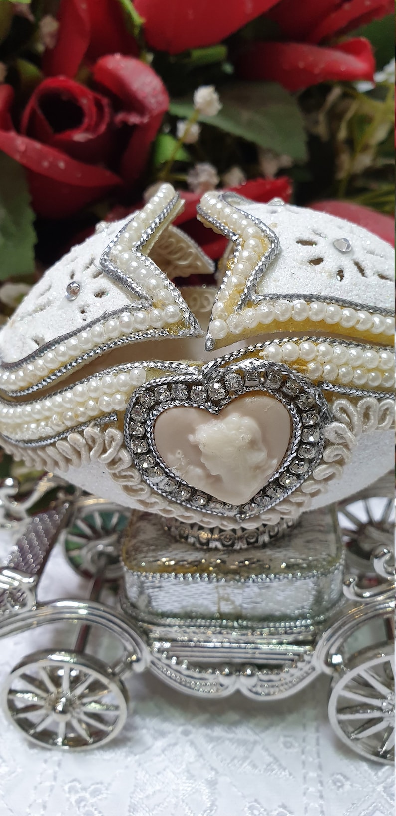 May include: A white porcelain egg decorated with pearls and rhinestones, shaped like a carriage with a heart-shaped cameo on the front. The carriage has silver wheels and a silver base.