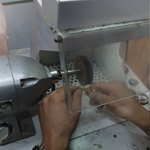 May include: A person is using a polishing machine to polish a gold piece of jewelry. The machine has a buffing wheel and a clear plastic shield to protect the user from dust and debris. The person is wearing a watch with a black and blue striped band.