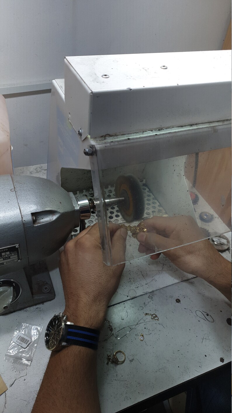 May include: A person is using a polishing machine to polish a gold piece of jewelry. The machine has a buffing wheel and a clear plastic shield to protect the user from dust and debris. The person is wearing a watch with a black and blue striped band.
