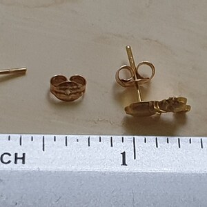 May include: A pair of gold-colored earrings with a small, detailed design. The earrings are shown next to a ruler, with the design measuring approximately 0.5 inches in length.