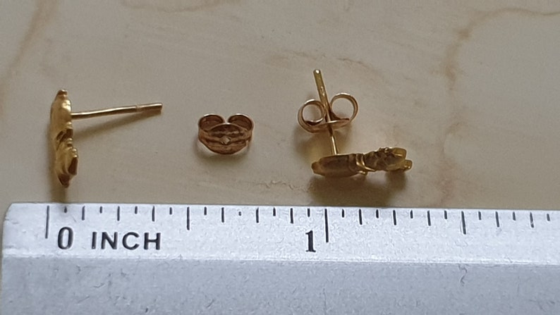 May include: A pair of gold-colored earrings with a small, detailed design. The earrings are shown next to a ruler, with the design measuring approximately 0.5 inches in length.