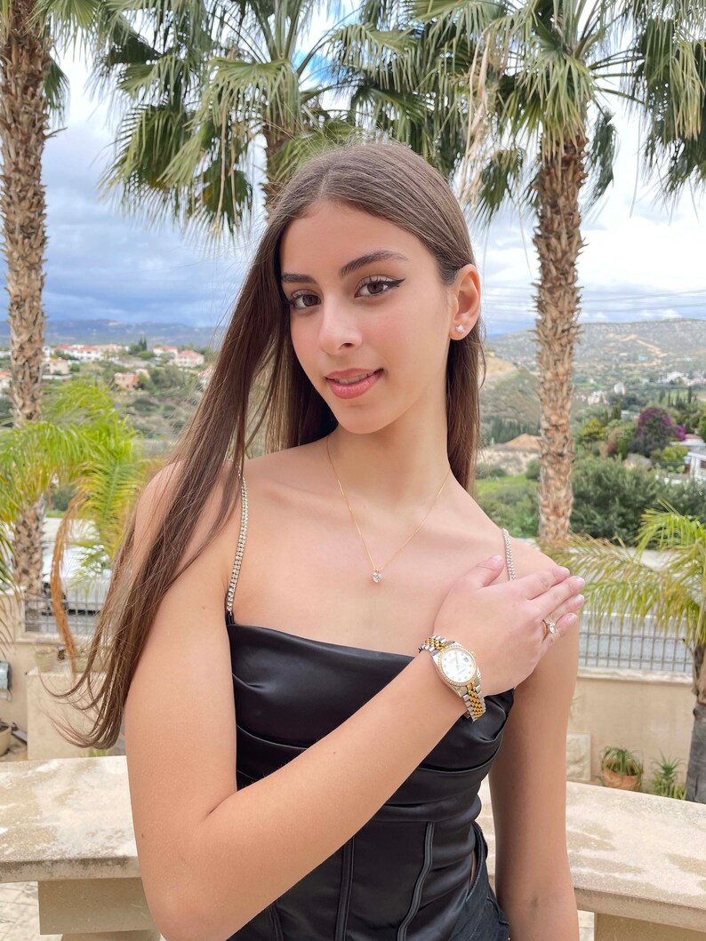 Pu&ograve; includere: Una donna indossa un top nero senza maniche con un orologio d'oro e argento al polso. &Egrave; in piedi davanti a una palma e ha i capelli lunghi e castani.