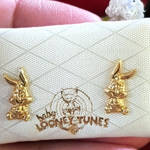May include: A white box with a gold Bugs Bunny design. The box has the words "Baby Looney Tunes" printed on it. The box contains a pair of gold Bugs Bunny earrings.