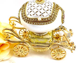 May include: A white porcelain egg shaped music box in the form of a golden carriage. The carriage has intricate details and is decorated with rhinestones. The egg is decorated with a diamond pattern and has a pearl on top.