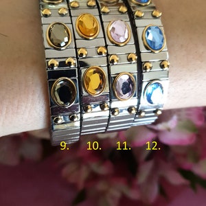 May include: Four silver and gold bracelets with different coloured gemstones. The bracelets are stacked on a person's wrist. The gemstones are blue, pink, yellow, and black.