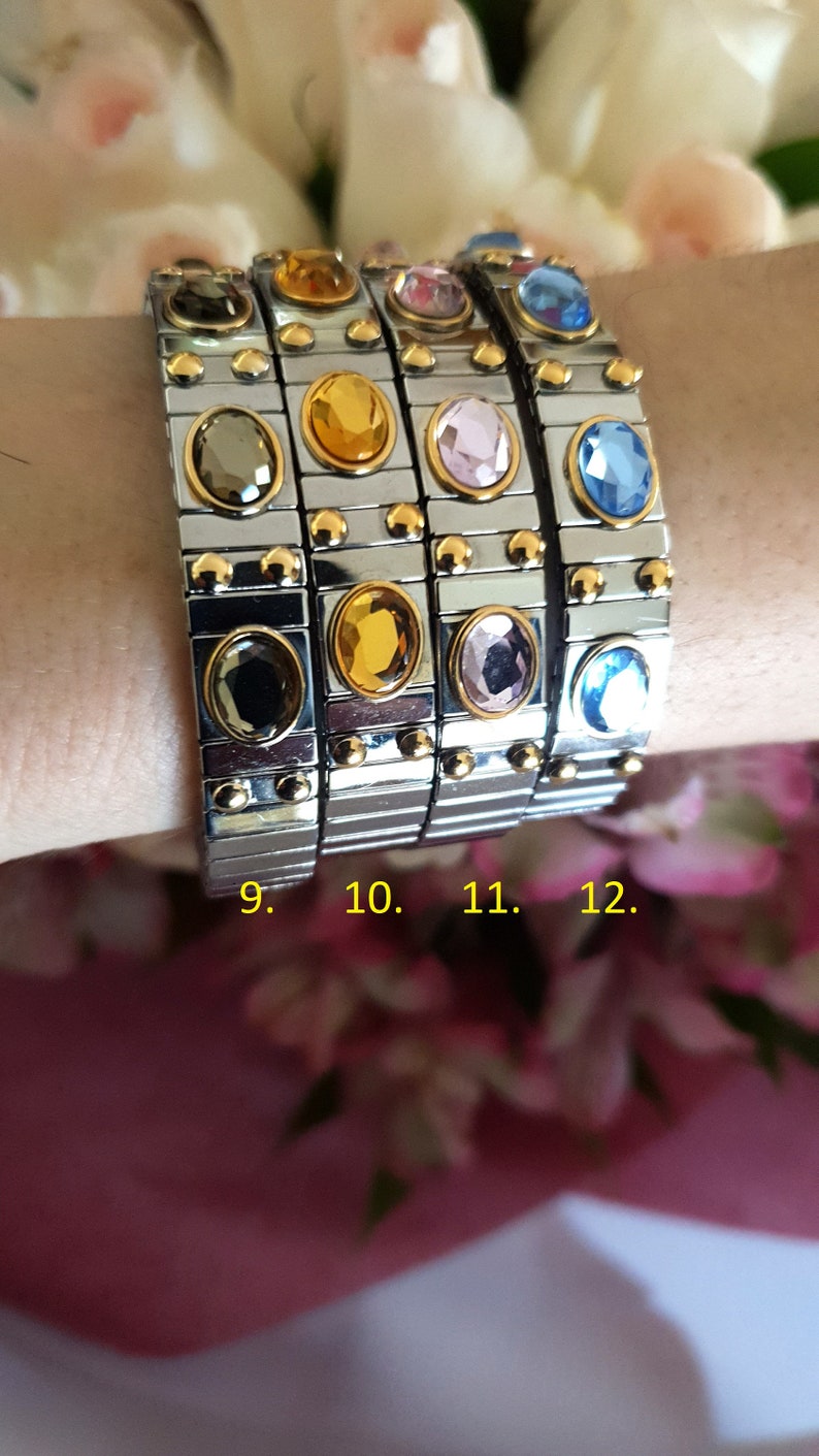 May include: Four silver and gold bracelets with different coloured gemstones. The bracelets are stacked on a person's wrist. The gemstones are blue, pink, yellow, and black.
