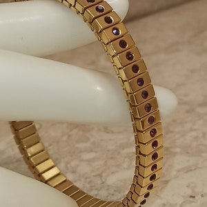 May include: A gold-toned bracelet with a flexible design. The bracelet is adorned with a row of small, dark purple gemstones.