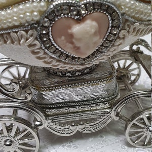May include: A white porcelain egg decorated with pearls and rhinestones, opened to reveal a pink heart-shaped cameo inside. The egg is mounted on a silver carriage with wheels.