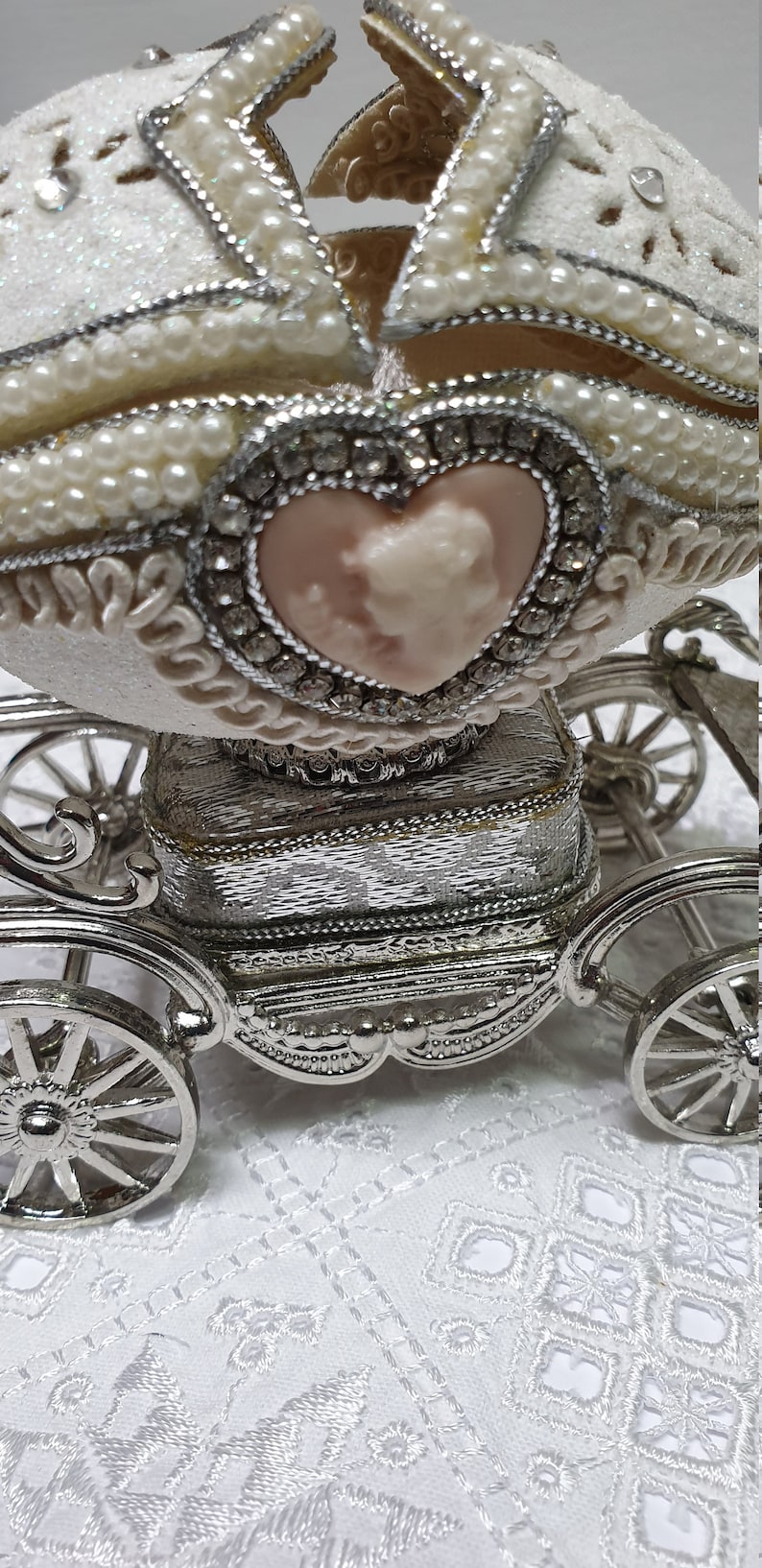 May include: A white porcelain egg decorated with pearls and rhinestones, opened to reveal a pink heart-shaped cameo inside. The egg is mounted on a silver carriage with wheels.