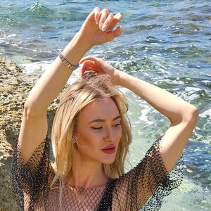 May include: A woman with long blonde hair wearing a black mesh top with a black bra underneath. She is standing in the water with her arms raised above her head.