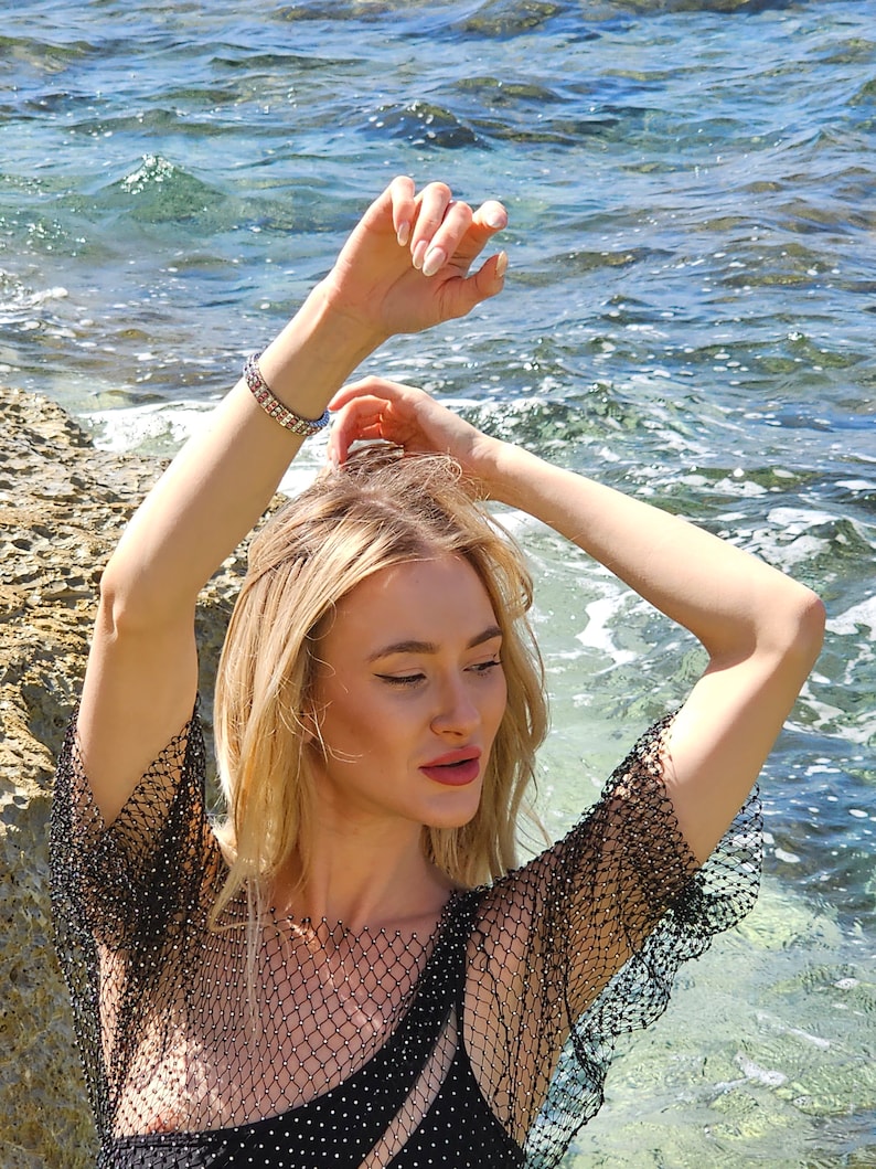 May include: A woman with long blonde hair wearing a black mesh top with a black bra underneath. She is standing in the water with her arms raised above her head.