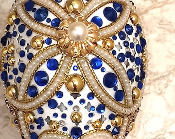 May include: A white egg decorated with blue and gold beads, pearls, and a floral design.