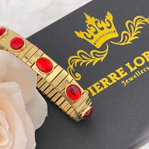 Puede incluir: Un brazalete magnético dorado con piedras ovaladas rojas. El brazalete está sobre una caja negra con el texto "PIERRE LORREN Joyeros".