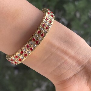 Puede incluir: Un brazalete dorado con cristales rojos y transparentes. El brazalete está hecho de pequeños eslabones cuadrados.