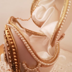 May include: A white porcelain egg with gold and pearl accents, opened to reveal a white satin interior. The egg is on a gold stand.