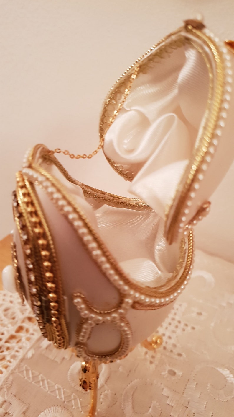 May include: A white porcelain egg with gold and pearl accents, opened to reveal a white satin interior. The egg is on a gold stand.