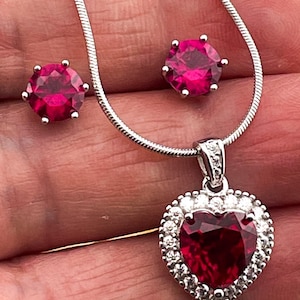 May include: A silver necklace with a heart-shaped pendant featuring a large red gemstone surrounded by smaller white gemstones. The necklace comes with a pair of matching red gemstone earrings.