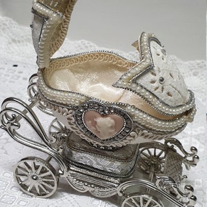 May include: A silver carriage-shaped trinket box with a white egg-shaped lid decorated with pearls and a pink cameo. The lid is open, revealing a white satin interior.