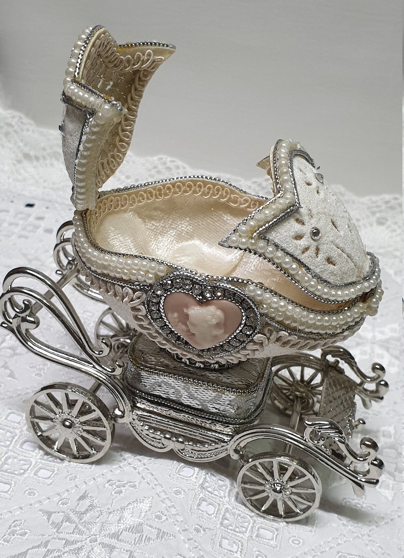 May include: A silver carriage-shaped trinket box with a white egg-shaped lid decorated with pearls and a pink cameo. The lid is open, revealing a white satin interior.