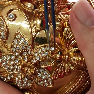 Puede incluir: Un adorno en forma de huevo de color dorado con detalles intrincados y un dise&ntilde;o de mariposa. El adorno est&aacute; cubierto de diamantes de imitaci&oacute;n transparentes y tiene un peque&ntilde;o dise&ntilde;o de abeja en el lateral. El adorno est&aacute; siendo sujetado por unas pinzas.
