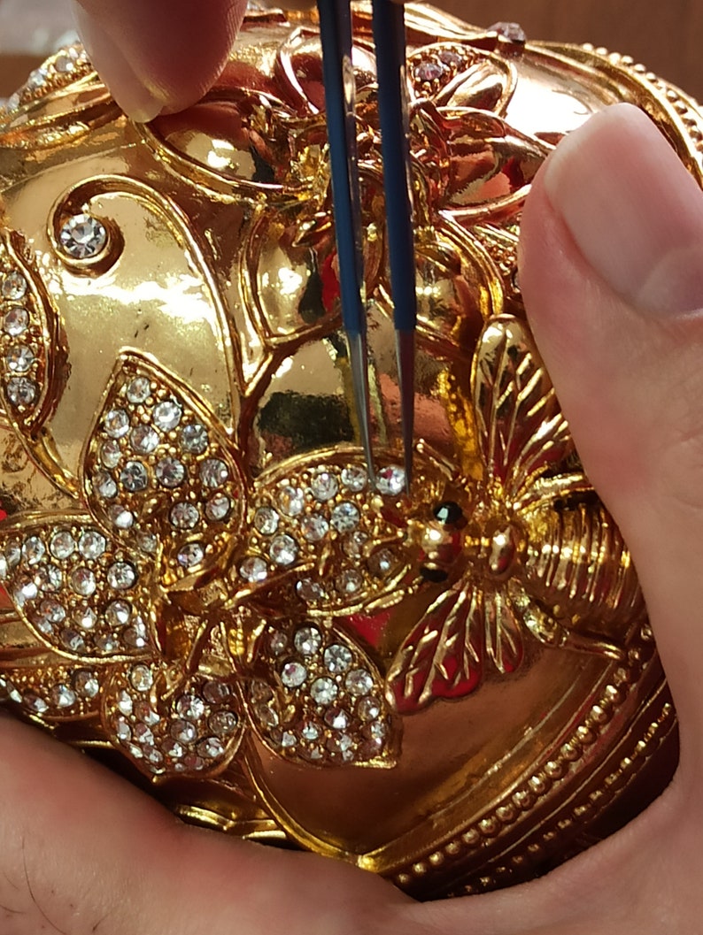 Puede incluir: Un adorno en forma de huevo de color dorado con detalles intrincados y un dise&ntilde;o de mariposa. El adorno est&aacute; cubierto de diamantes de imitaci&oacute;n transparentes y tiene un peque&ntilde;o dise&ntilde;o de abeja en el lateral. El adorno est&aacute; siendo sujetado por unas pinzas.