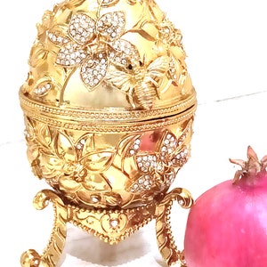 Puede incluir: Un huevo decorativo dorado con un dise&ntilde;o de abeja y flores, adornado con diamantes de imitaci&oacute;n, se encuentra sobre un soporte. Una granada rosa est&aacute; al lado del huevo.