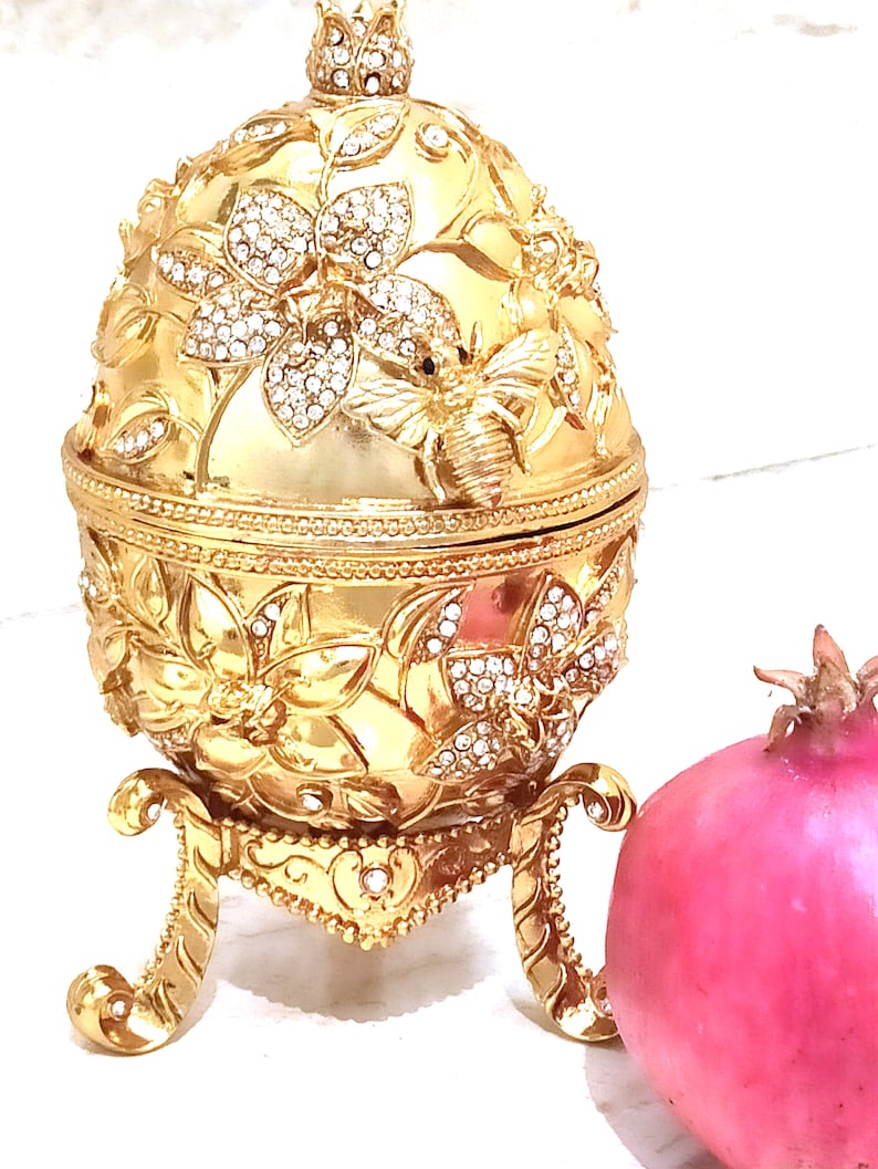 Puede incluir: Un huevo decorativo dorado con un dise&ntilde;o de abeja y flores, adornado con diamantes de imitaci&oacute;n, se encuentra sobre un soporte. Una granada rosa est&aacute; al lado del huevo.