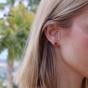 May include: A close-up of a person's face, with a focus on a small, red gemstone stud earring. The earring is set in a simple design. The person has blonde hair and is outdoors with a blurred green background.