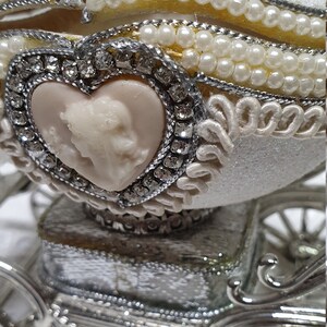 May include: A white porcelain egg decorated with pearls and rhinestones, depicting a carriage with a heart-shaped cameo. The egg is open and sits on a silver carriage base.