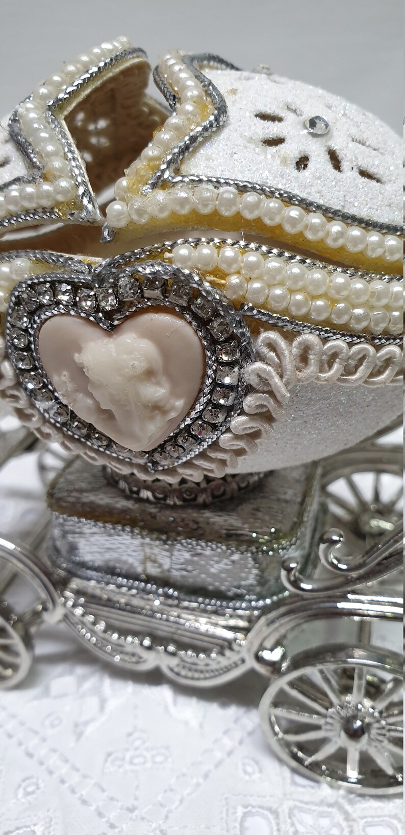 May include: A white porcelain egg decorated with pearls and rhinestones, depicting a carriage with a heart-shaped cameo. The egg is open and sits on a silver carriage base.