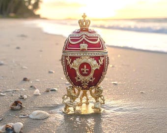 May include: A red and gold decorative egg with a crown on top, resting on a gold stand with lion figures. The egg is embellished with ornate gold detailing and jewels. The background shows a beach at sunset.