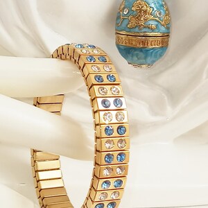 Puede incluir: Un brazalete dorado con cristales azules y transparentes. El brazalete est&aacute; hecho de cuadrados de metal unidos. El brazalete est&aacute; sobre una superficie blanca.