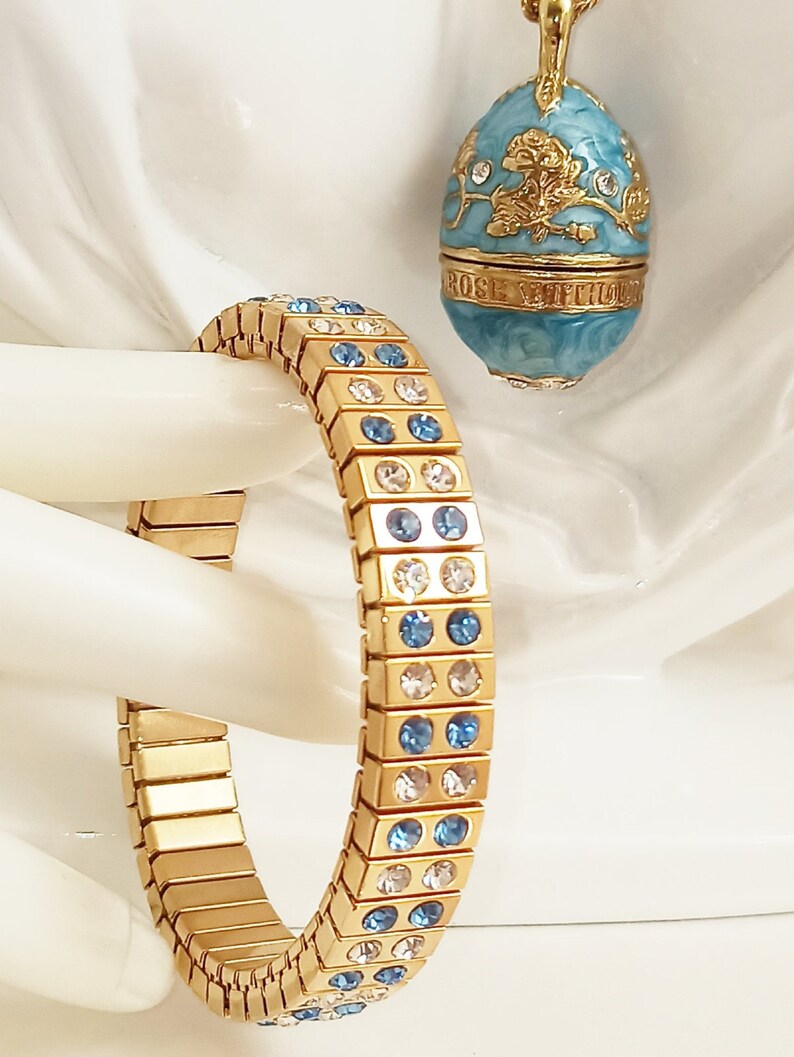 Puede incluir: Un brazalete dorado con cristales azules y transparentes. El brazalete est&aacute; hecho de cuadrados de metal unidos. El brazalete est&aacute; sobre una superficie blanca.