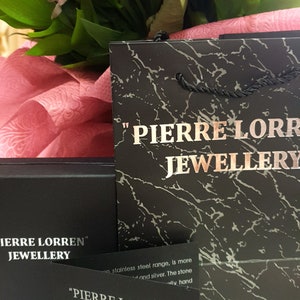 May include: A black gift bag with the text "PIERRE LORREN JEWELLERY" printed on it. The bag is sitting on a pink surface with a bouquet of pink flowers.