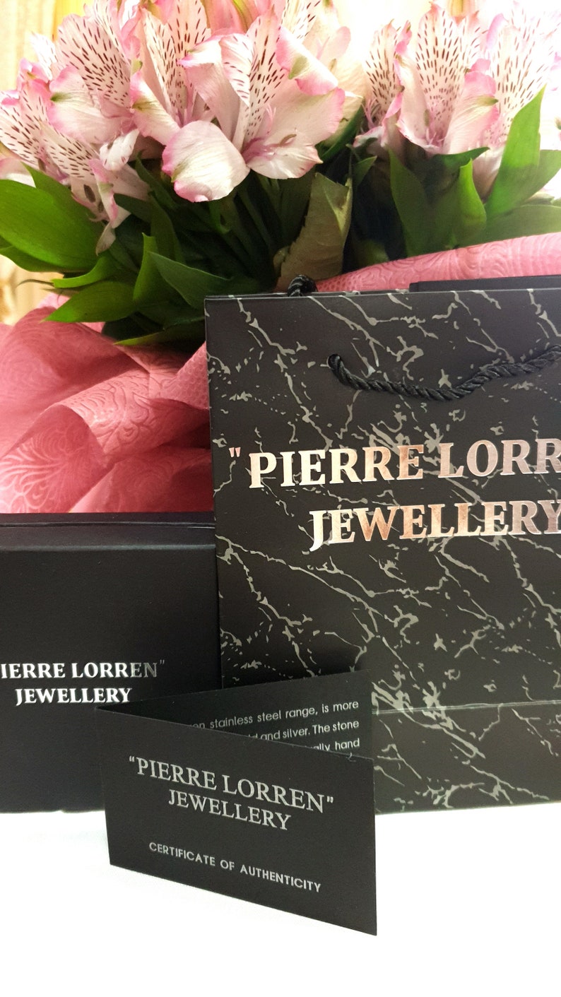 May include: A black gift bag with the text "PIERRE LORREN JEWELLERY" printed on it. The bag is sitting on a pink surface with a bouquet of pink flowers.