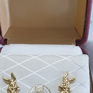 May include: A pink velvet jewelry box with a white interior. The box is open and shows two gold-colored Bugs Bunny earrings. The box has the words "18 CT GOLD" and "Looney Tunes" printed on it.