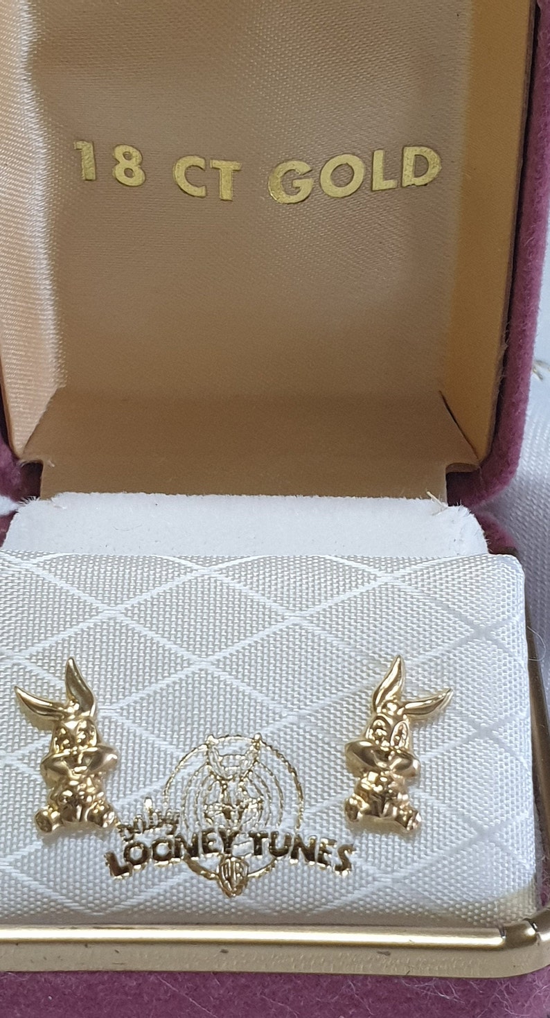 May include: A pink velvet jewelry box with a white interior. The box is open and shows two gold-colored Bugs Bunny earrings. The box has the words "18 CT GOLD" and "Looney Tunes" printed on it.