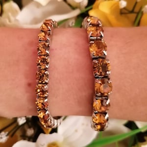 May include: Two silver bracelets with amber-colored crystal stones. The bracelets are on a person's wrist.