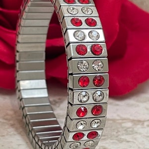 May include: A silver expandable bracelet with red and clear crystals. The bracelet is made of metal links and has a modern design.