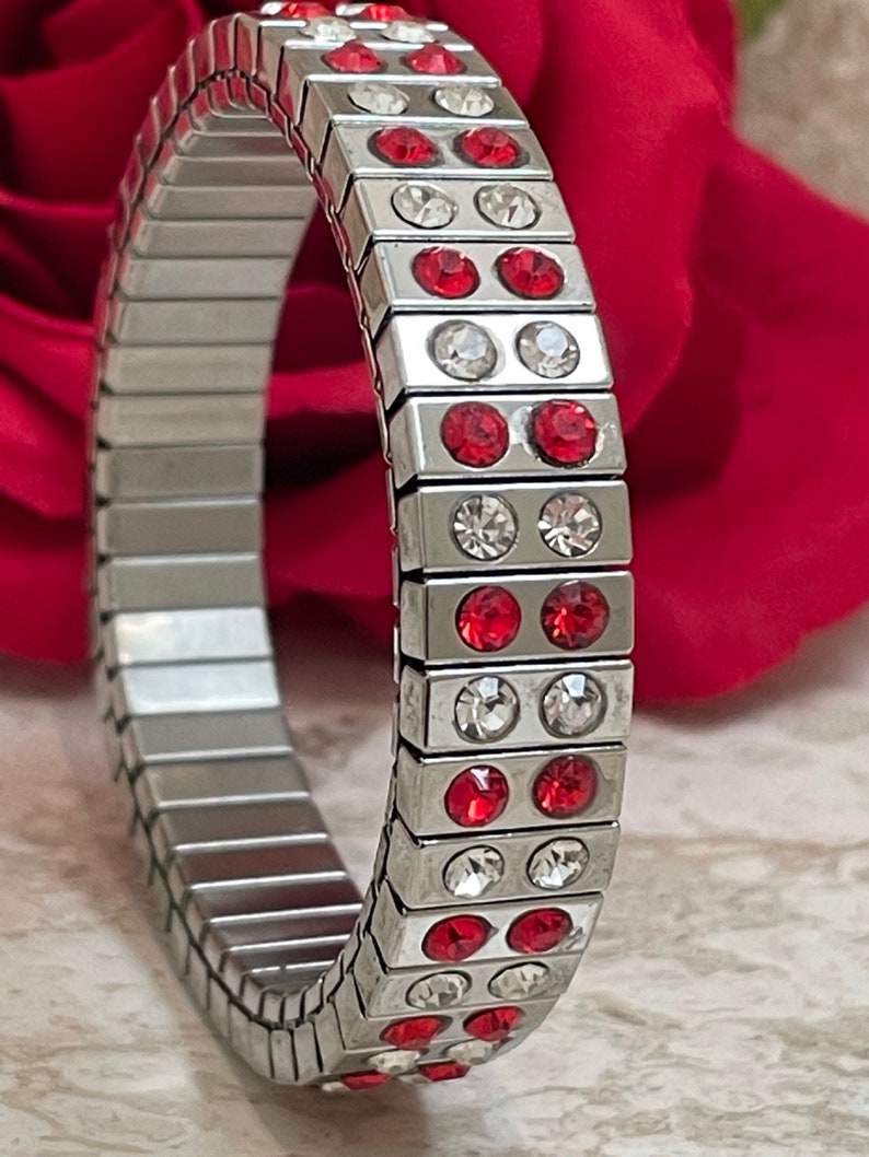 May include: A silver expandable bracelet with red and clear crystals. The bracelet is made of metal links and has a modern design.
