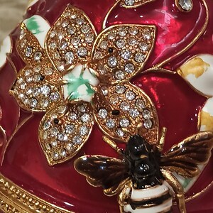 May include: A red, gold and white jeweled egg with a bee and flower design. The egg is decorated with rhinestones and has a gold trim.