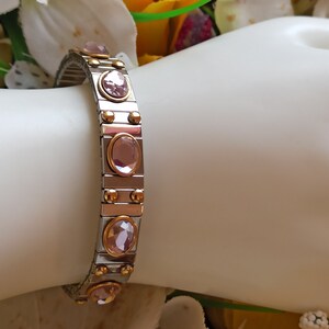 May include: A silver and gold bracelet with a row of pink oval-shaped stones. The bracelet is made of metal links and has a magnetic clasp.